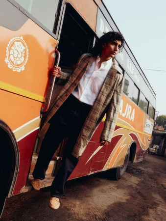 A young adult man in a plaid jacket and white shirt stands in the doorway of an orange bus on a street in the daytime.