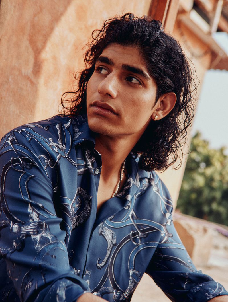Young man with long dark curly hair wearing a patterned blue button-down shirt outdoors against textured terracotta walls.