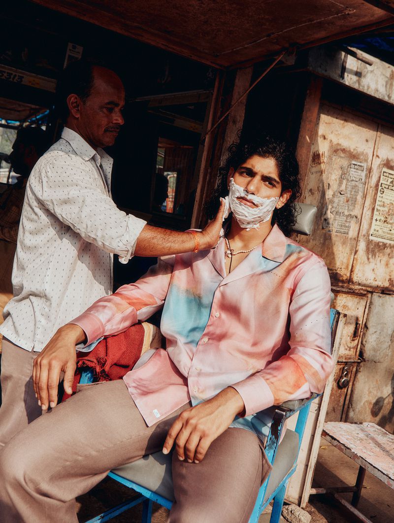 Adult male seated in a chair outdoors getting a shave from another adult male barber in a brightly lit outdoor environment.