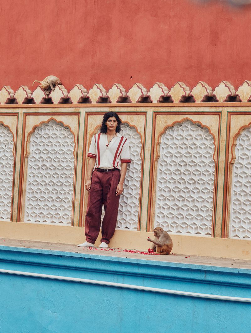 Adult male poses next to monkey on outdoor set with vivid peach wall in the background, wearing casual top and slacks. Lifestyle fashion.