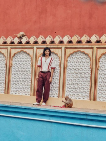 Adult male poses next to monkey on outdoor set with vivid peach wall in the background, wearing casual top and slacks. Lifestyle fashion.