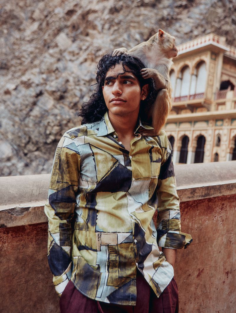 Young adult male with long dark hair in patterned shirt poses with a macaque monkey on his shoulders in an outdoor setting.