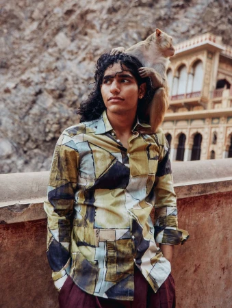 Young adult male with long dark hair in patterned shirt poses with a macaque monkey on his shoulders in an outdoor setting.