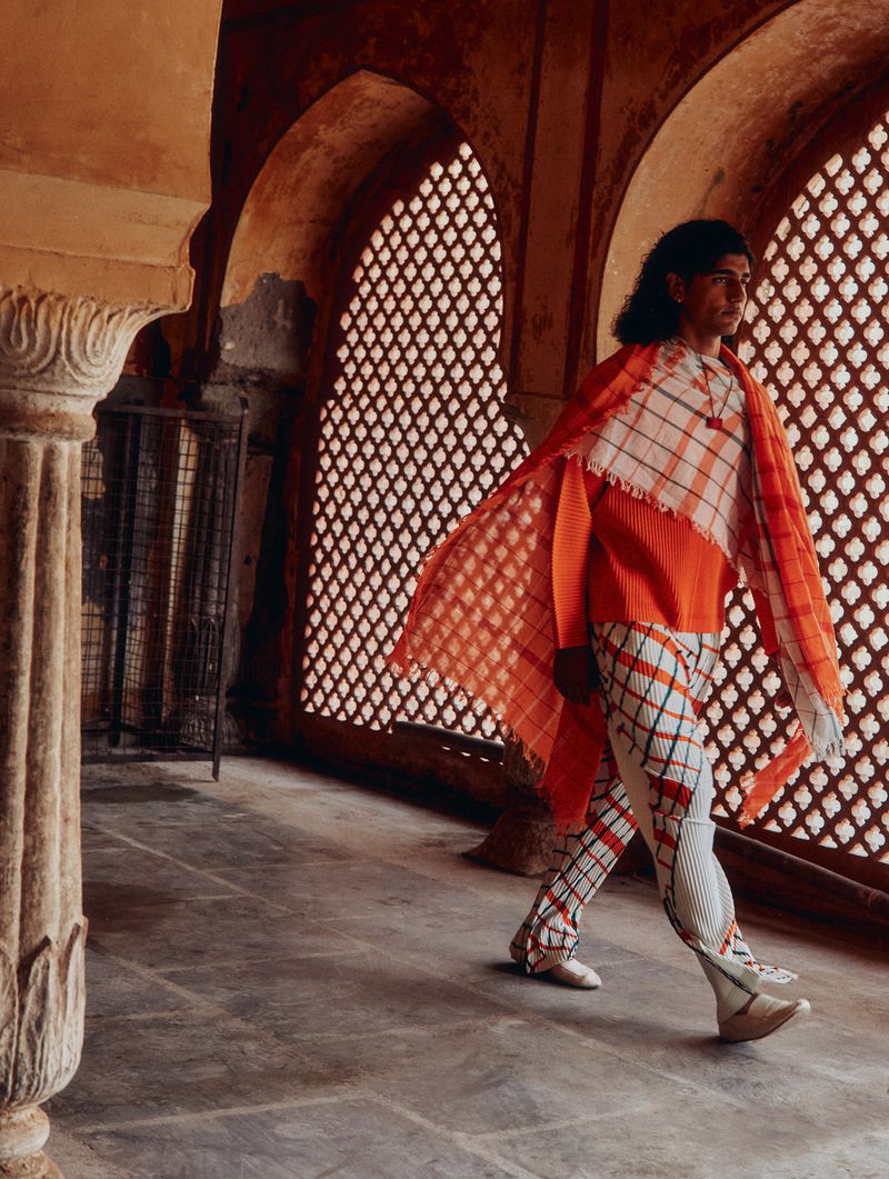 A fashion shot of an adult male in a pleated orange top, patterned scarf and pants walking through a columned interior space with latticed windows.