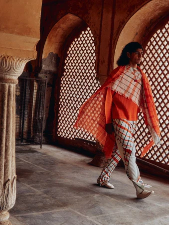 A fashion shot of an adult male in a pleated orange top, patterned scarf and pants walking through a columned interior space with latticed windows.