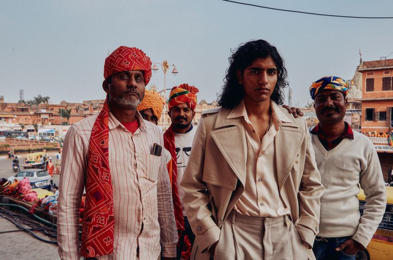 Group portrait featuring a fashionably dressed person in a beige trench coat surrounded by individuals in traditional Indian attire outdoors.