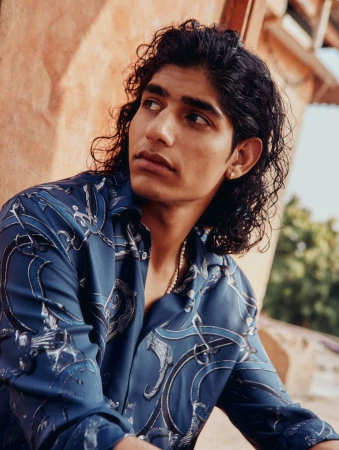 Young man with long dark curly hair wearing a patterned blue button-down shirt outdoors against textured terracotta walls.
