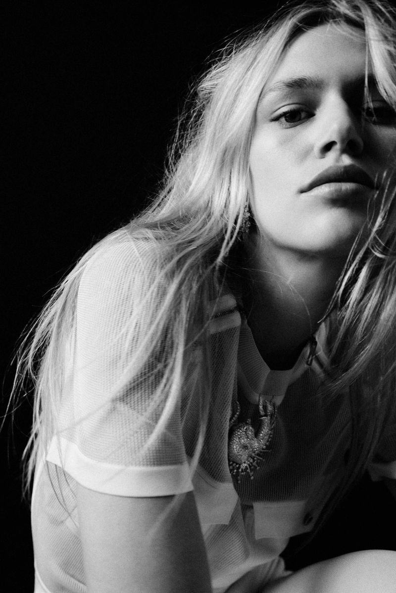 Monochrome portrait of a young woman with long blonde hair, wearing a white mesh top, against a solid black background.