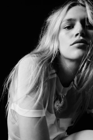 Monochrome portrait of a young woman with long blonde hair, wearing a white mesh top, against a solid black background.