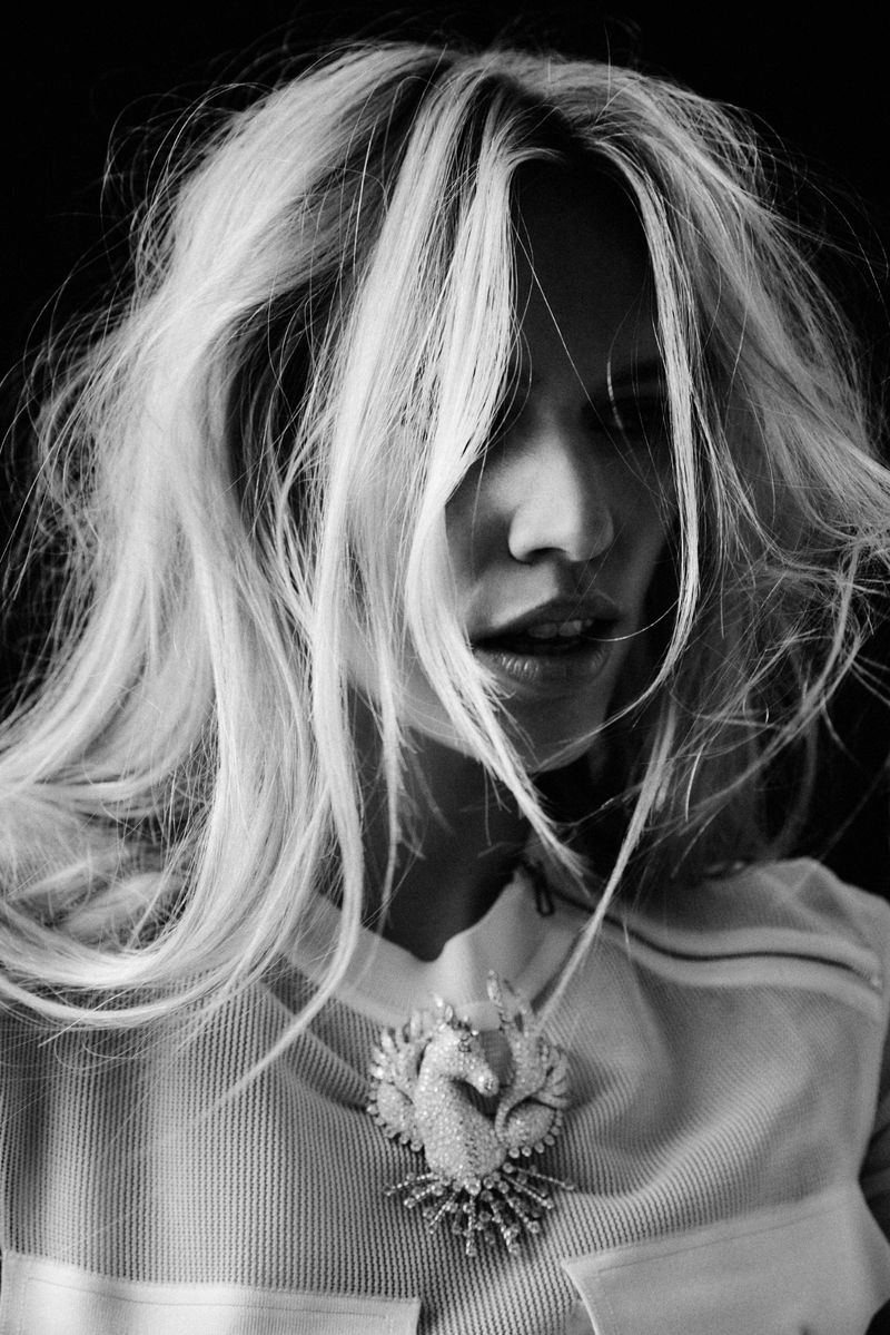 Monochrome portrait of a woman with wavy blond hair wearing a white shirt and embellished brooch against a black background.