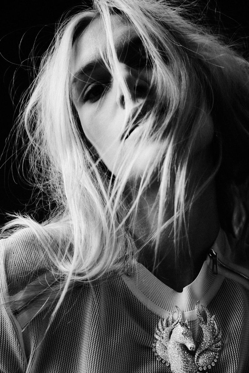 Black and white portrait of a woman with long blonde hair blowing around her face, wearing a light-colored top with a unicorn brooch.