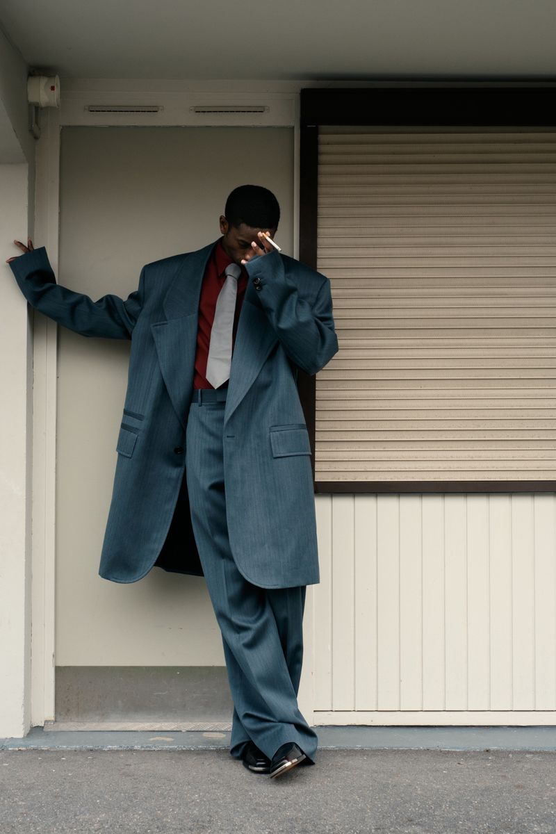 Man in oversized teal suit posing against wall with closed shutter, wearing red shirt and grey tie, urban setting with neutral tones.