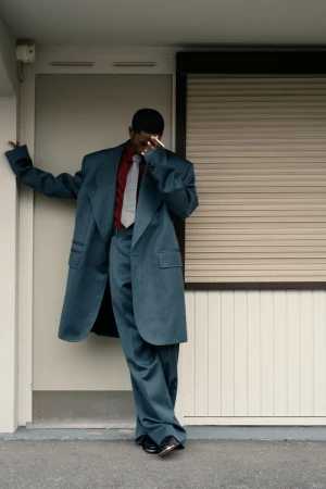 Man in oversized teal suit posing against wall with closed shutter, wearing red shirt and grey tie, urban setting with neutral tones.