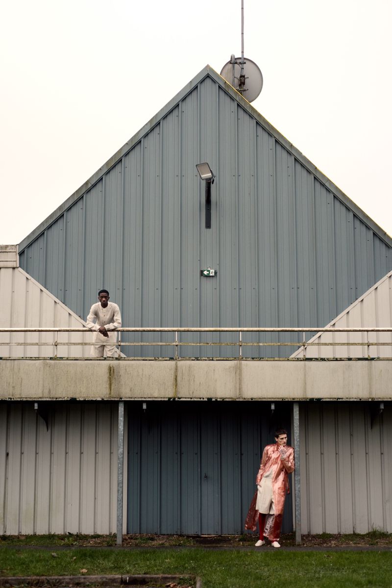 Two people stand outside an institutional building with metal siding; one on the balcony and one on the ground; overcast day.