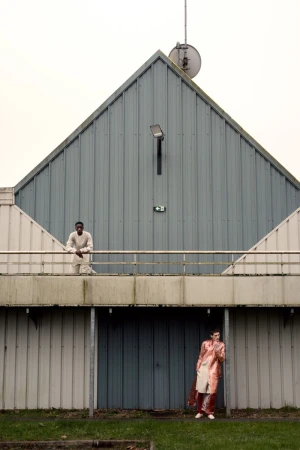 Two people stand outside an institutional building with metal siding; one on the balcony and one on the ground; overcast day.