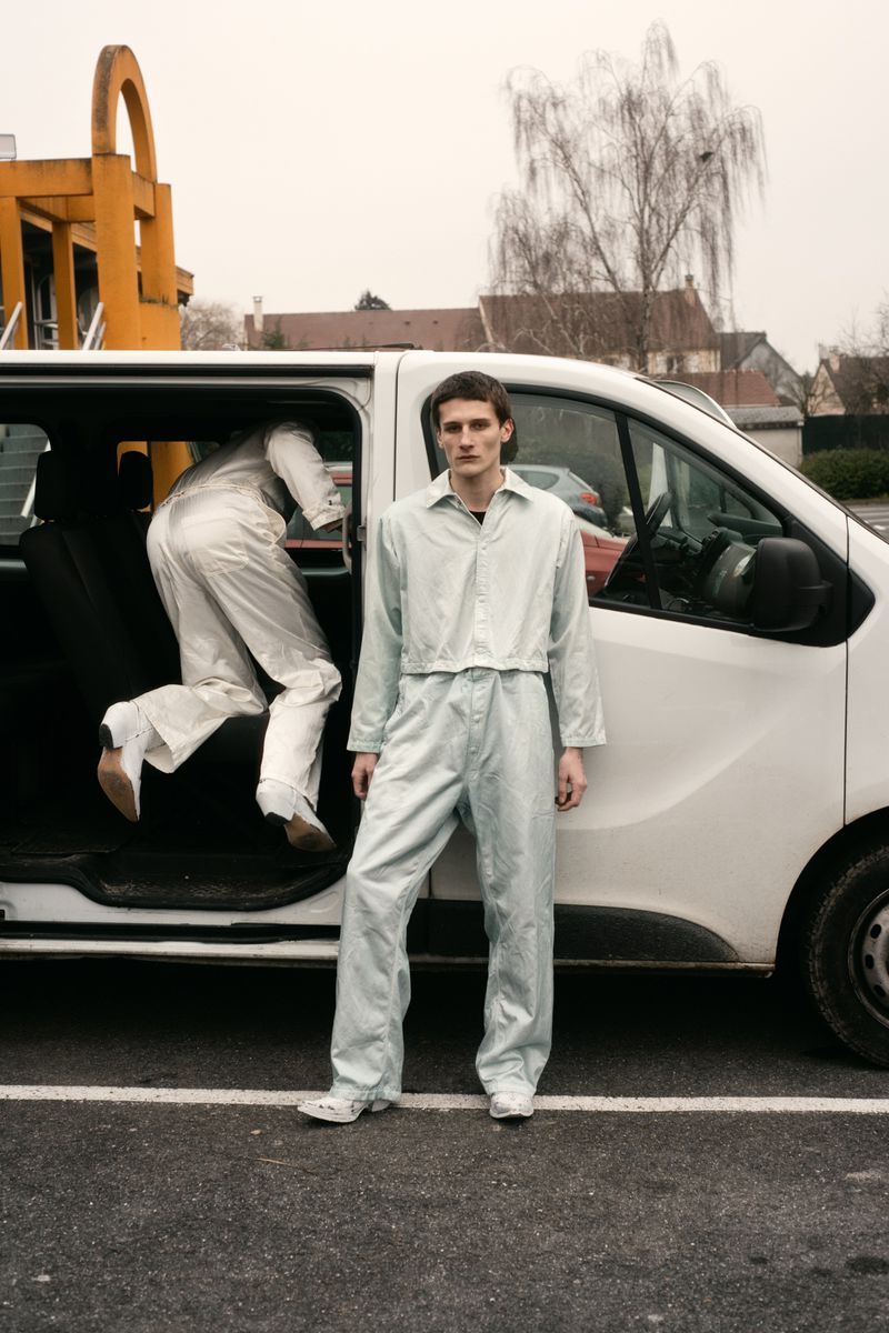 Two adult males in matching mint green outfits near a white van in an urban parking lot on a cloudy day.