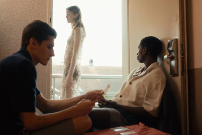 Indoor scene features three people: one walking and two seated. One seated is playing cards, and a closed door is to the right.