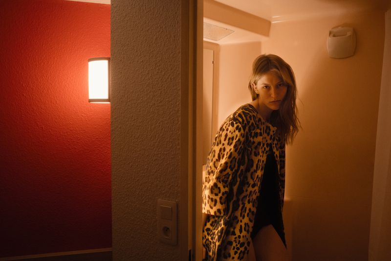 A woman with long hair gazes at the camera, wearing a leopard print coat. Interior shot with reddish wall and doorway in soft, diffused light.