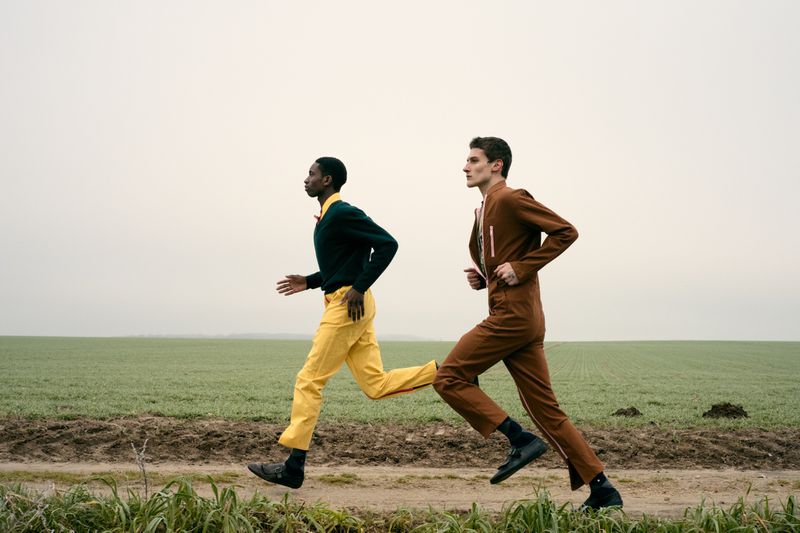 Two adult models running on a dirt path through a field under an overcast sky. One man wears yellow pants and the other a brown jumpsuit.