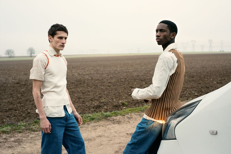 Two young men in vintage-inspired outfits stand near a white car on a dirt road, set against a rural landscape under an overcast sky.