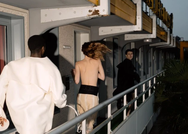 Three people run on the walkway of an apartment complex on a dark day, two shirtless, wearing minimal clothing and one in a white jacket.