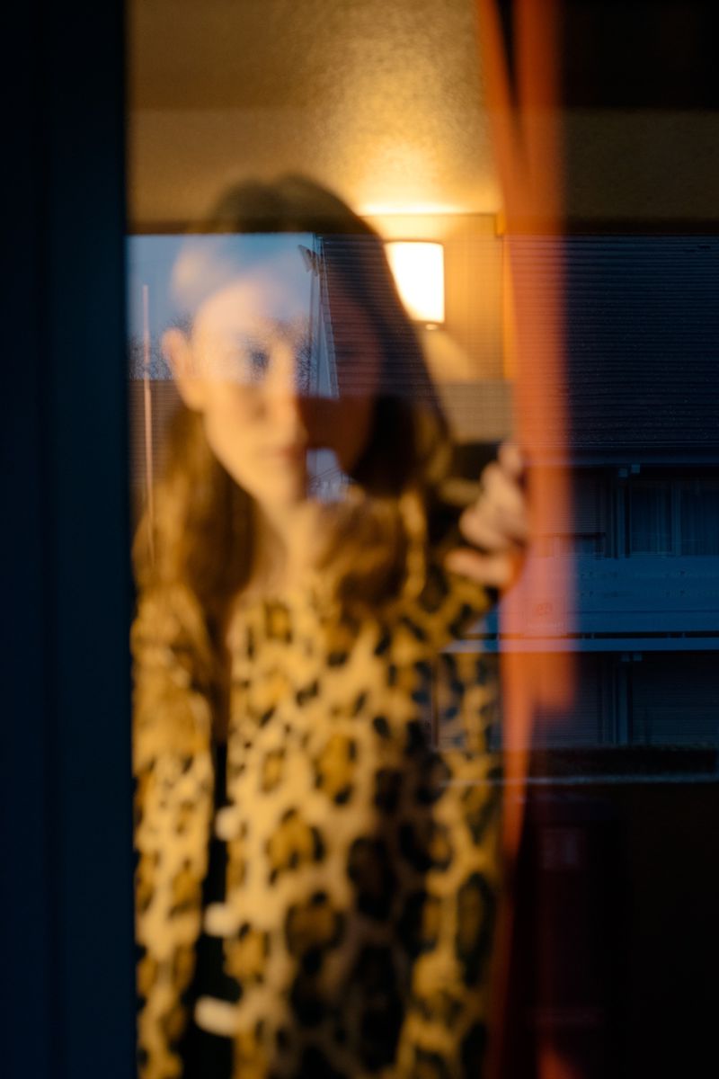 Portrait of a woman with long hair wearing an animal-print jacket, seen through a window with reflections of interior lighting.