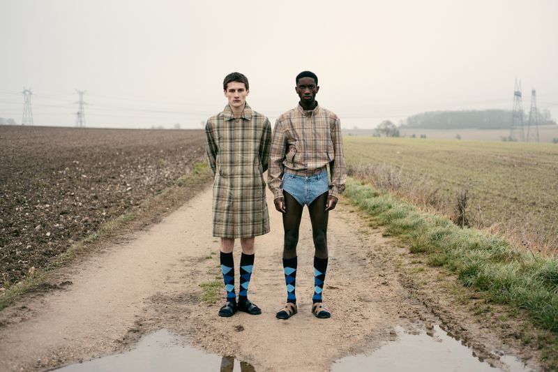 Two people stand on a dirt road in a field; one is wearing a plaid coat, and the other is wearing a matching shirt and jean shorts.
