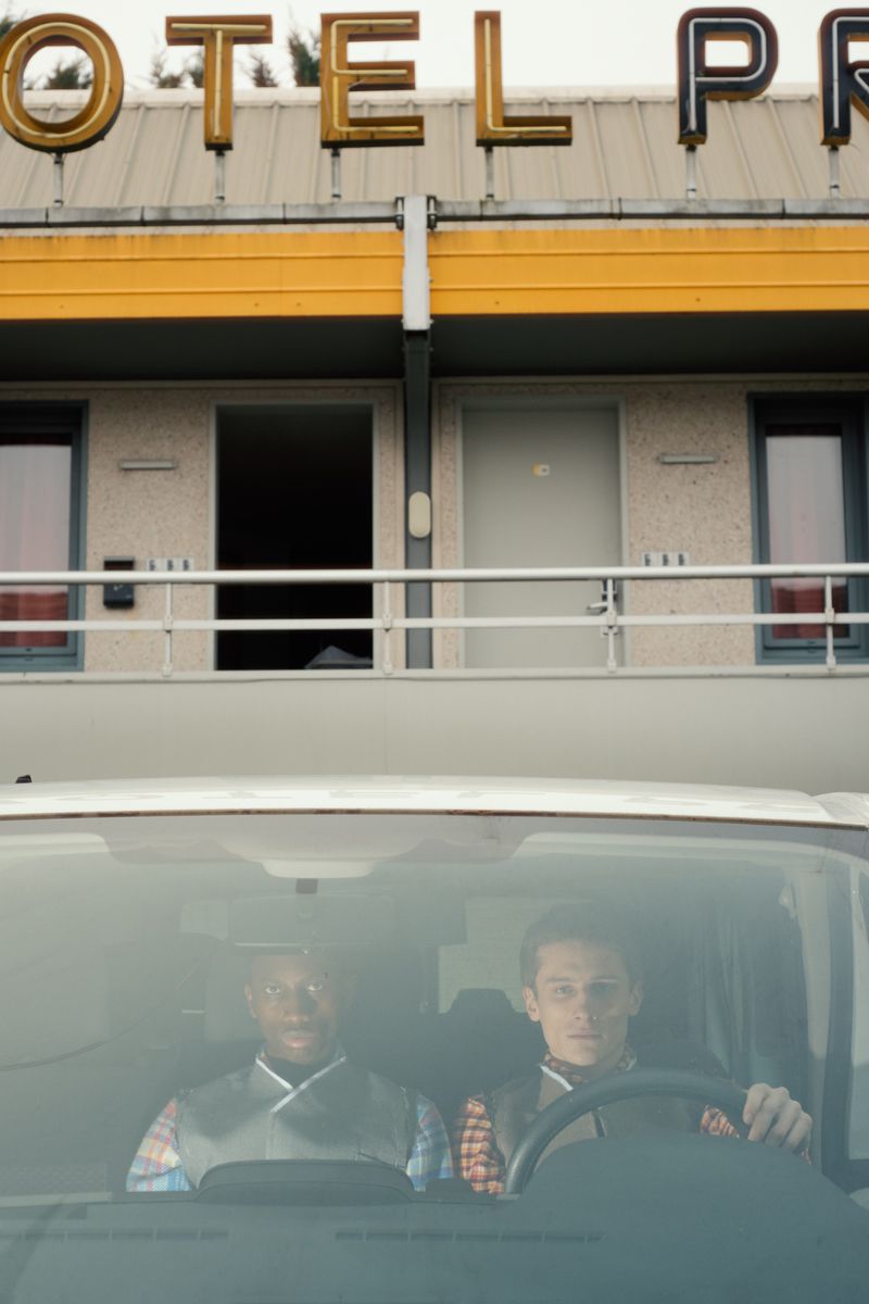 Two men sit inside a car parked in front of a vintage motel with a yellow sign. The composition captures an urban setting.