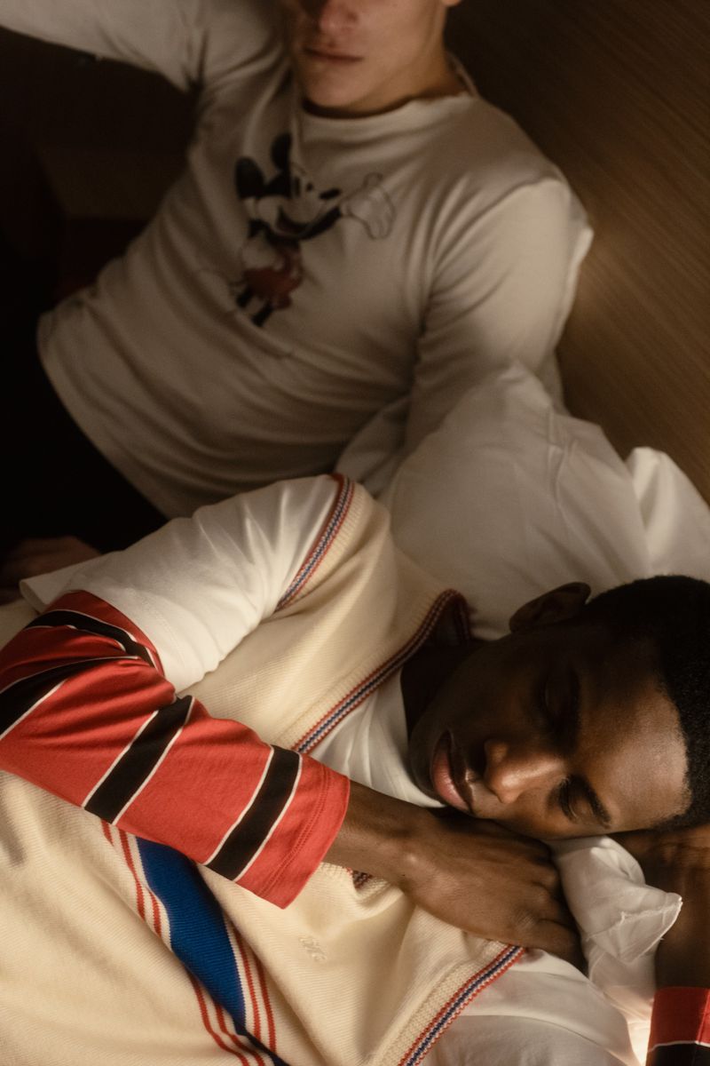Two men in a bedroom. One is light-skinned and sitting up. The other is dark-skinned, lying down, and appears to be sleeping in the indoors setting.