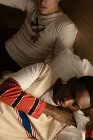 Two men in a bedroom. One is light-skinned and sitting up. The other is dark-skinned, lying down, and appears to be sleeping in the indoors setting.