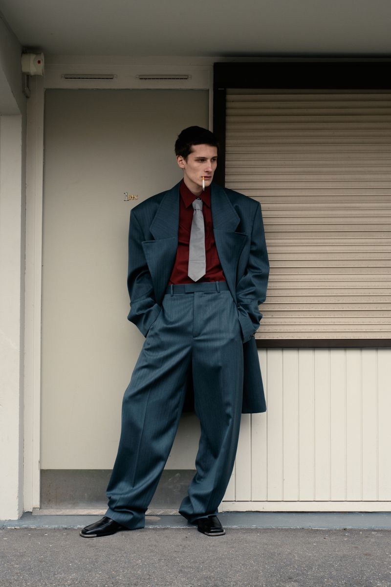 Stylish man in oversized teal suit and red shirt stands against neutral wall with cigarette in mouth, in a medium shot.