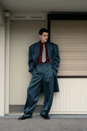 Young man in an oversized teal suit with a dark red shirt and grey tie leaning against a doorway