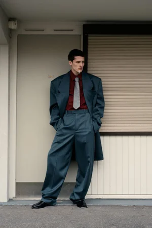 Young man in an oversized teal suit with a dark red shirt and grey tie leaning against a doorway