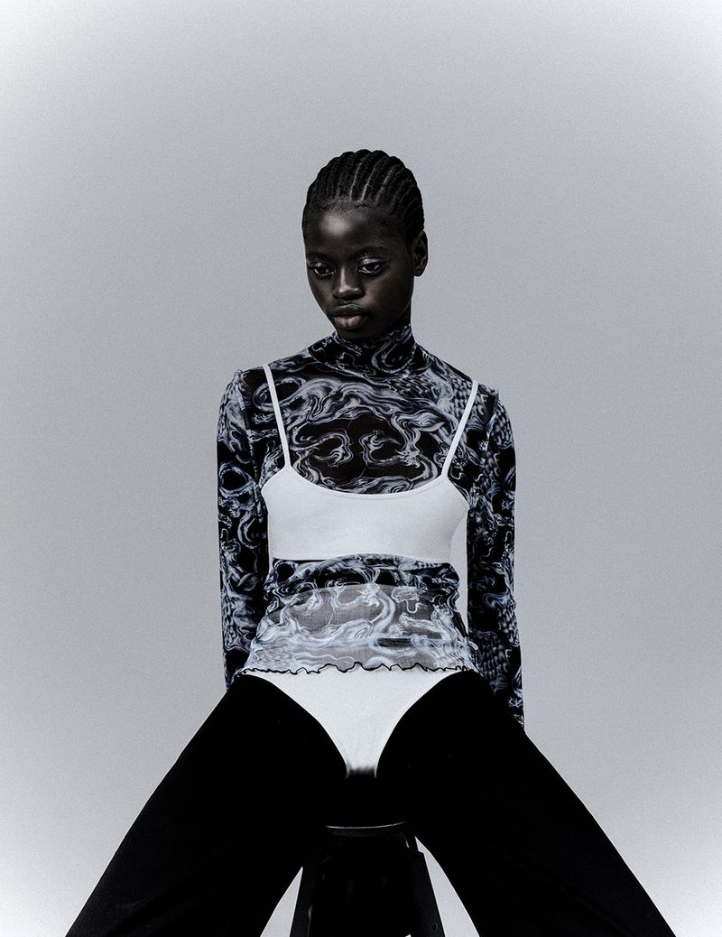 A dark-skinned woman with braided hair sits in a studio wearing a patterned long-sleeved top, white cropped tank, and black pants.