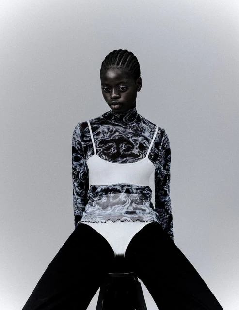 A dark-skinned woman with braided hair sits in a studio wearing a patterned long-sleeved top, white cropped tank, and black pants.