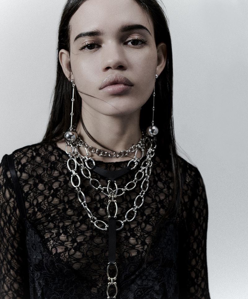 Portrait of a woman with long dark hair wearing a black lace top and layered silver necklaces and earrings against a white background.