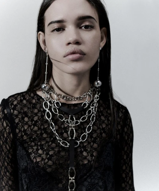 Portrait of a woman with long dark hair wearing a black lace top and layered silver necklaces and earrings against a white background.