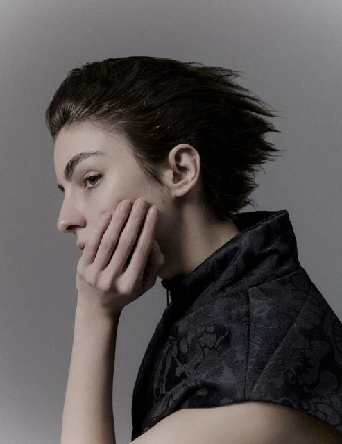 Profile portrait of a woman with dark hair, gazing left while resting her cheek in her hand, wearing a patterned, high-collared top.