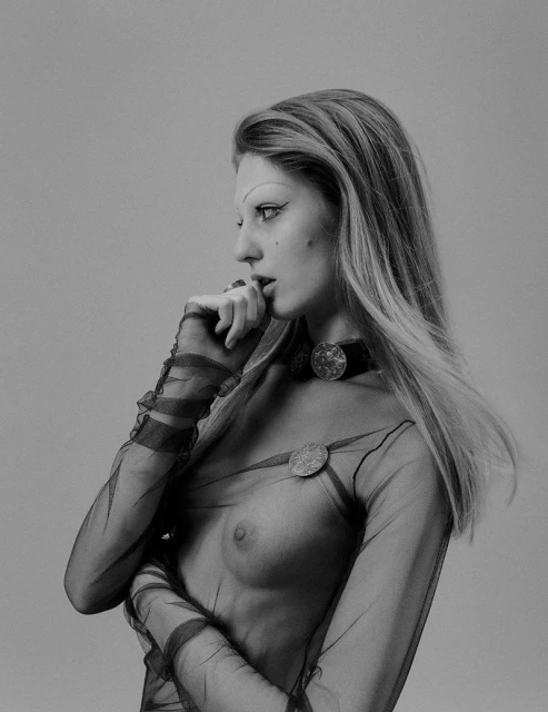 Striking black-and-white portrait of a young woman with long blonde hair, wearing a sheer top and unique choker in a neutral studio.