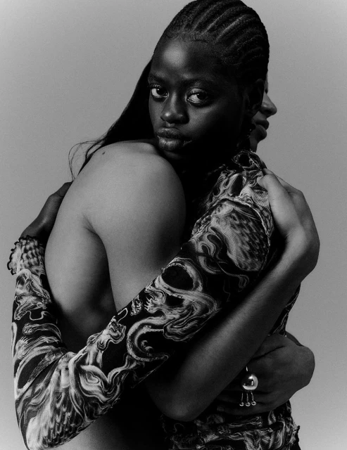 Monochrome portrait of two women hugging in patterned tops, one looking directly at the viewer with long dark hair and braided hair.