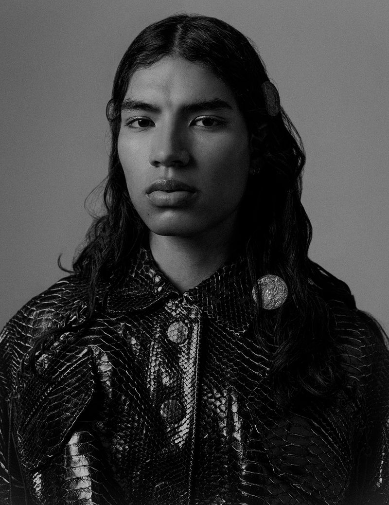 Portrait of an adult with long hair wearing a textured jacket and round hair clips against a neutral grey studio backdrop.