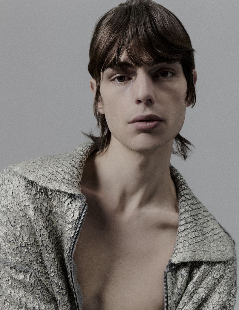 Portrait of a young adult male in a textured silvery-grey jacket, indoors against a grey backdrop with diffused lighting. Fashion editorial.
