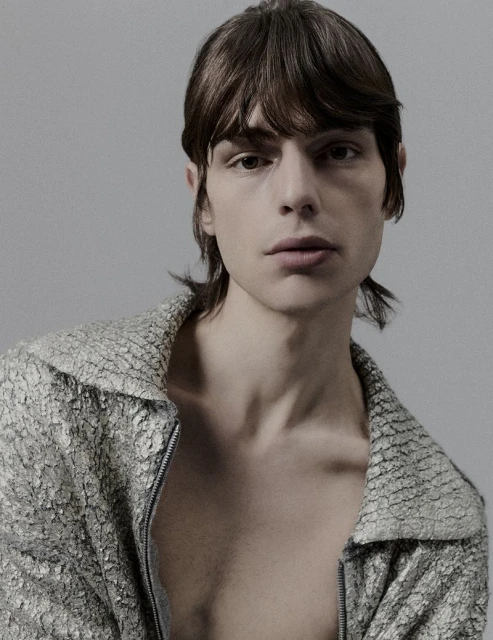 Portrait of a young adult male in a textured silvery-grey jacket, indoors against a grey backdrop with diffused lighting. Fashion editorial.