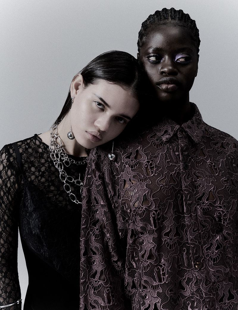 Two fashion models in a studio shot, one wearing a black lace dress and silver jewelry, the other wearing a patterned button-down shirt.