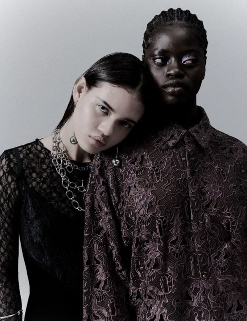 Two fashion models in a studio shot, one wearing a black lace dress and silver jewelry, the other wearing a patterned button-down shirt.