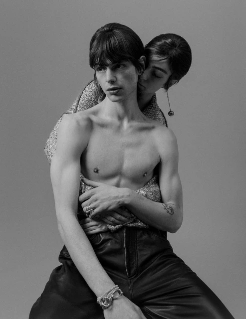 Two young adults in a close embrace, the person in front is shirtless wearing leather pants, the person behind wears shiny top.