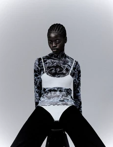 Black model wearing patterned sheer turtleneck and white slip top against plain grey studio background