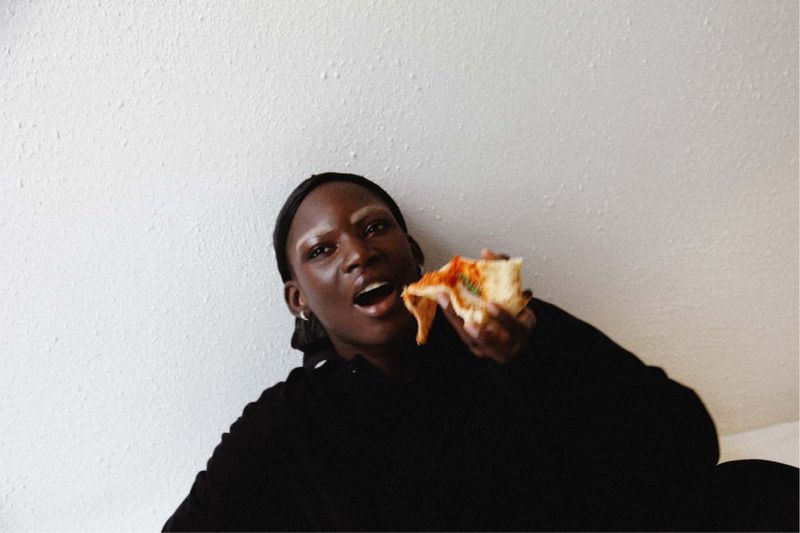 A young African-descent woman with a dark sweater is joyfully biting into a slice of pizza against a textured white backdrop.