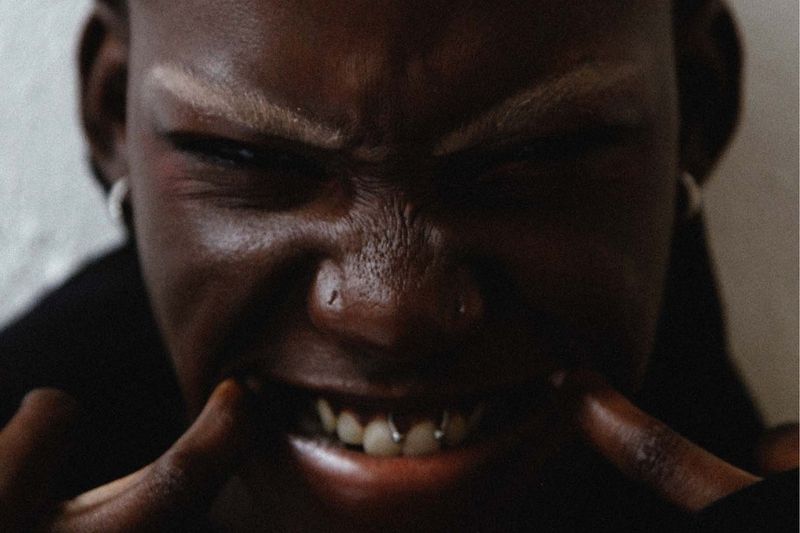 Close-up of a young adult with dark skin, fair eyebrows, lip piercings, and metal teeth, pulling on their lips with their fingers.