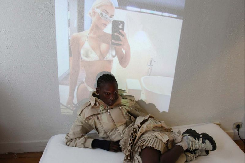 Woman in avant-garde khaki outfit reclines in a white room; a projection of a bikini-clad woman with sunglasses appears on the wall behind.